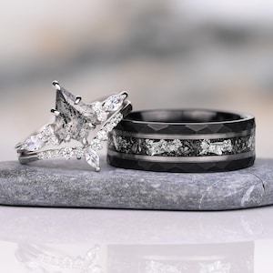 Puede incluir: Un conjunto de anillos plateados con un diamante sal y pimienta en forma de cometa, acentuado por piedras blancas redondas y marquesas, junto a una banda negra facetada con una incrustación central de material plateado triturado.