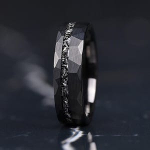 Peut inclure: Une bague noire présentant une surface extérieure facettée et martelée avec une incrustation centrale en matériau métallique texturé. L'anneau possède une finition mate sombre aux bords géométriques nets et un canal central argenté.
