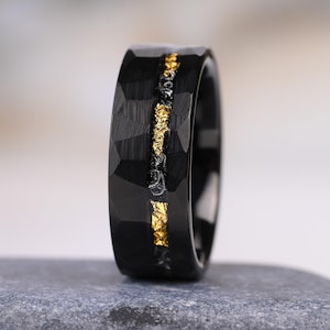 May include: A black ring with a faceted, hammered exterior surface. A central horizontal inlay runs around the circumference, featuring a textured, dark gray base embedded with irregular, shimmering gold-colored metallic flakes.