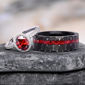 Puede incluir: Un anillo de compromiso de plata con una piedra roja de corte ovalado rodeada por un halo de pequeñas piedras blancas, junto a una banda negra ancha con textura martillada e incrustación central de material rojo.