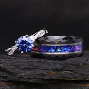 Puede incluir: Un anillo plateado con una piedra azul redonda de talla brillante y banda con pequeñas piedras claras, junto a un anillo negro facetado con una incrustación central de material azul y morado que recuerda a una galaxia.