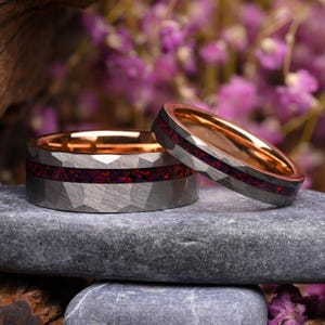 Pode incluir: Dois anéis com exterior de metal cinzento martelado e interior de ouro rosa polido. Cada banda apresenta uma incrustação central de pedra vermelha escura triturada ou material de opala que percorre horizontalmente a circunferência.