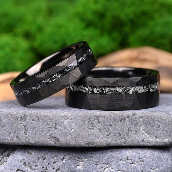 May include: Two black rings with a faceted exterior surface and a central inlay featuring a textured, metallic silver-toned material. The rings have a dark, matte finish and a geometric, multi-sided design around the outer band circumference.
