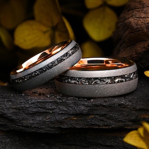Könnte beinhalten: Zwei Ringe mit einer Innenseite aus Roségold und einem texturierten grauen Außenband. Ein mittiger Einsatz aus dunklem, fragmentiertem Material verläuft horizontal um den Umfang jedes Rings und bildet einen Kontrast zum matten grauen Metall.