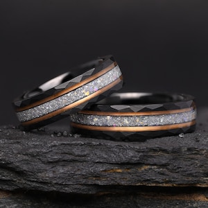 Könnte beinhalten: Zwei schwarze Ringe mit facettierter Außenfläche. Jeder Ring verfügt über eine mittige Einlage aus schimmerndem, hellem, zerkleinertem Material, die von dünnen, metallischen, kupferfarbenen Bändern entlang des Umfangs eingefasst ist.