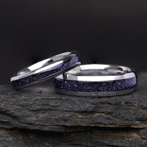 Puede incluir: Dos anillos de tono plateado con una incrustación central de material azul oscuro brillante. Los anillos tienen un acabado metálico pulido y reflectante con un perfil de banda redondeado, mostrando una textura azul profunda y centelleante.