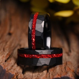 Könnte beinhalten: Ein schwarzer Ring mit gehämmertem Muster und einer zentralen Einlage aus zerkleinertem rotem Material. Das Band hat eine dunkle, matte Oberfläche mit geometrischen Facetten, die durch einen leuchtenden, roten horizontalen Streifen ergänzt wird.