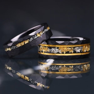 May include: Two black rings with a faceted exterior surface. Each ring features a central inlay containing gold-colored flakes and silver-colored metallic fragments. The ring on the right is bordered by two thin, smooth gold-colored bands.