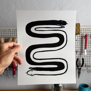 May include: A rectangular white paper print featuring a black and white illustration of two snakes intertwined in a long, S-shaped pattern. The top snake is black with a white outline, and the bottom snake is white with a black outline.