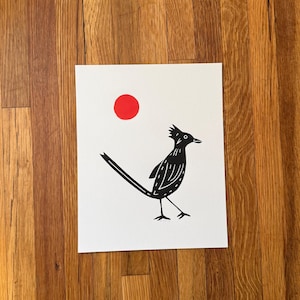 May include: A rectangular white art print featuring a black ink illustration of a roadrunner bird with a crest on its head and a long tail. Above the bird is a solid red circle. The bird has stylized white line details within its black body.