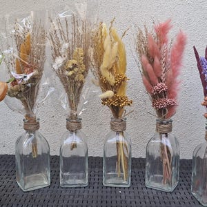 May include: Six clear glass bottles with twine-wrapped necks, each containing a unique arrangement of dried flowers and grasses in various colors including white, yellow, pink, and deep purple, displayed in a row against a neutral wall.