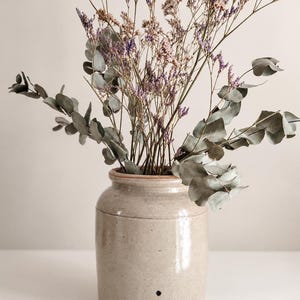 Peut inclure: Un pot en céramique beige de forme cylindrique contenant un bouquet de branches d'eucalyptus séchées aux feuilles gris-vert arrondies et de longues tiges fines de petites fleurs de statice violettes et rose pâle, avec un petit point noir.
