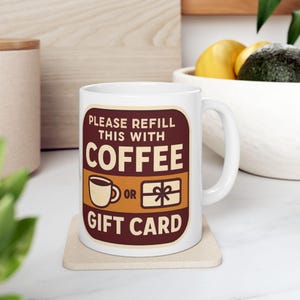 May include: A white ceramic mug featuring a dark brown rectangular graphic with rounded corners. The text reads: PLEASE REFILL THIS WITH COFFEE OR GIFT CARD. The design includes a small illustration of a coffee cup and a wrapped gift box.