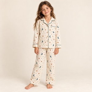 Può includere: Un set pigiama a due pezzi in tessuto color crema con un motivo ripetuto di lune crescenti blu ottanio scuro e stelle a quattro punte color ruggine. La camicia a maniche lunghe abbottonata presenta un colletto e un taschino, abbinata a pantaloni lunghi coordinati.