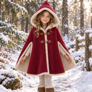 May include: A deep red hooded cape made of textured fleece material with a cream-colored sherpa lining. The garment features two brown toggle closures on the front and a pointed hood, providing a layered look over a light-colored long-sleeve top.