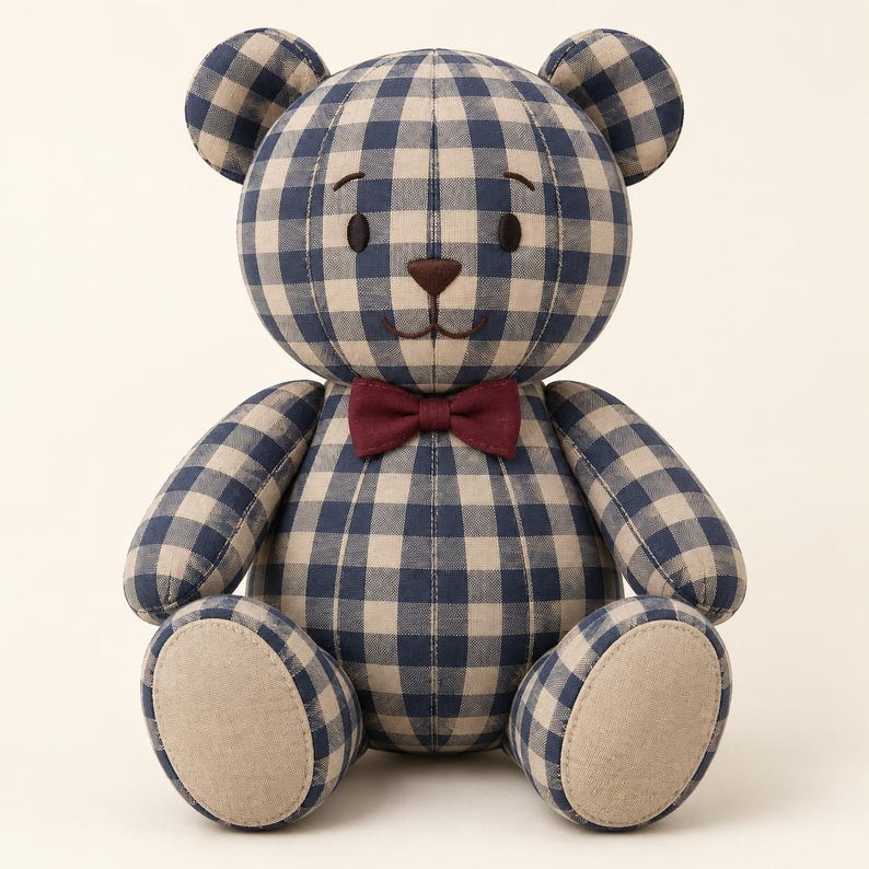 Puede incluir: Un oso de peluche con un patr&oacute;n de cuadros vichy en azul marino y beige en el cuerpo, la cabeza y las extremidades. El oso tiene ojos bordados en marr&oacute;n oscuro, una nariz marr&oacute;n, una boca cosida y una pajarita roja oscura lisa en el cuello.