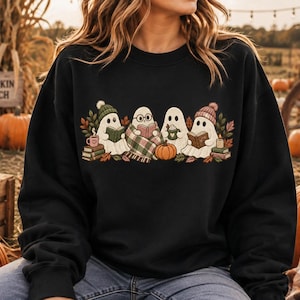 May include: A black long-sleeve sweatshirt featuring a graphic print of four white ghosts. The ghosts are wearing knit hats, reading books, and holding mugs, surrounded by autumn leaves, pumpkins, and stacks of books in shades of orange and green.