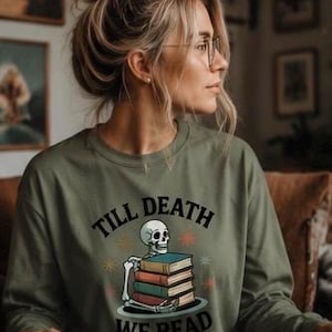 May include: An olive green long-sleeve crewneck sweatshirt featuring a graphic print of a skeleton resting its arms on a stack of four colourful books. The text "TILL DEATH WE READ" is printed in black capital letters above and below the illustration.