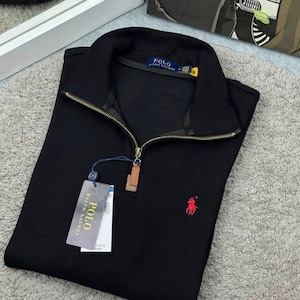 May include: A black long-sleeve pullover with a quarter-zip brass-toned metal closure, a high collar, and a red embroidered horse logo on the chest. The garment features a blue interior neck label reading Polo Ralph Lauren and a hanging retail tag.