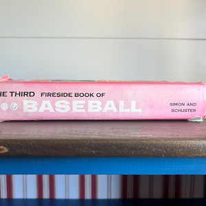 Könnte beinhalten: Ein gebundenes Buch mit dem Titel The Third Fireside Book of Baseball von Simon and Schuster. Der Buchrücken ist leuchtend rosa mit weißem Text und kleinen weißen Baseball-Grafiken. Das Buch weist Gebrauchsspuren an den Kanten und Ecken auf.