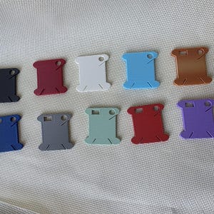 May include: Ten plastic embroidery floss bobbins arranged in two rows. The bobbins are rectangular with notched sides and a circular hole at the top, featuring a variety of solid colors including black, maroon, white, light blue, brown, blue, gray, sage green, red, and purple.