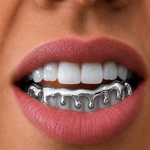 Puede incluir: Una pieza de grillz plateada colocada sobre los dientes inferiores, con un diseño distintivo de goteo líquido que se extiende desde la línea de las encías. El accesorio metálico tiene una superficie lisa y reflectante que se ajusta.