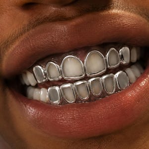 May include: A set of silver-coloured metal teeth grills covering the upper and lower teeth. The grills feature individual polished frames that outline each tooth, creating a metallic border around the natural white enamel of the teeth.