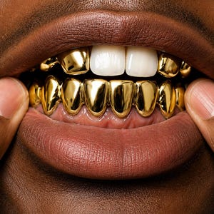 May include: A set of gold teeth grills covering the lower teeth, featuring a polished, reflective metallic finish. The upper teeth are natural white, with two gold caps visible on the upper left side, contrasting with the dark skin tone.