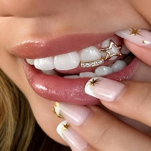 Peut inclure: Un bijou dentaire en or composé d'un contour en forme d'étoile et d'une barre horizontale sertie de petites pierres claires. Il est placé sur une dent, assorti à des ongles ornés d'étoiles dorées sur une base rose pâle et blanche.