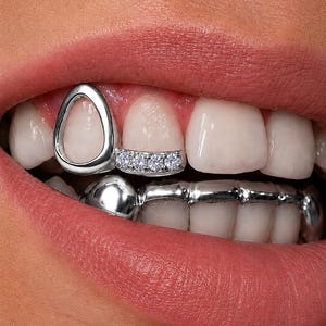 Puede incluir: Un grill dental de plata que presenta un marco en forma de lágrima en un diente superior y una barra horizontal con cuatro pequeñas piedras redondas transparentes en el diente adyacente, junto a un grill de plata liso que cubre los dientes inferiores.