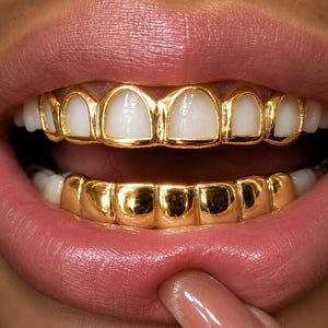 May include: A set of gold dental grills featuring a top row with individual gold frames around each white tooth and a solid, smooth, polished gold bottom row that covers the lower teeth completely, creating a reflective metallic appearance.