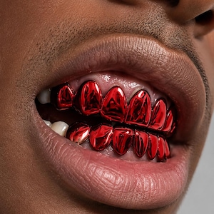 Op de afbeelding: Een set metallic rode tandgrillz die de boven- en ondertanden bedekken. De grillz hebben een sterk reflecterende, glanzende afwerking die de vorm van elke individuele tand volgt, wat zorgt voor een opvallende en levendige uitstraling.