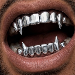 Könnte beinhalten: Ein Set aus silberfarbenen Metall-Zahngrillz, die die oberen und unteren Zähne bedecken. Das Design weist spitze, fangzahnartige Verlängerungen an den Eckzähnen beider Zahnreihen auf, was ein metallisches, reflektierendes Aussehen über den natürlichen Zähnen erzeugt.
