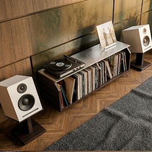 May include: Two white bookshelf speakers with black drivers, each mounted on a dark wooden stand. The speakers are positioned on either side of a low media console containing a black record player and a collection of vinyl records in sleeves.