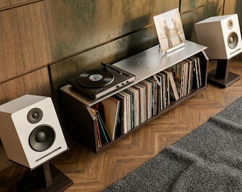 Op de afbeelding: Twee witte boekenplankluidsprekers met zwarte drivers, elk gemonteerd op een donkere houten standaard. De luidsprekers staan aan weerszijden van een laag mediameubel met een zwarte platenspeler en een verzameling vinylplaten in hoezen.