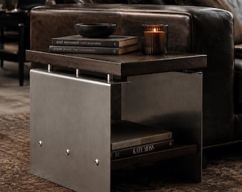 Op de afbeelding: Een bijzettafel met een donkerhouten blad en een onderste plank, ondersteund door twee geborstelde metalen zijpanelen die zijn vastgezet met zichtbare zilveren klinknagels. Op de tafel liggen twee boeken, een kleine donkere kom en een geribbelde glazen kaarshouder.