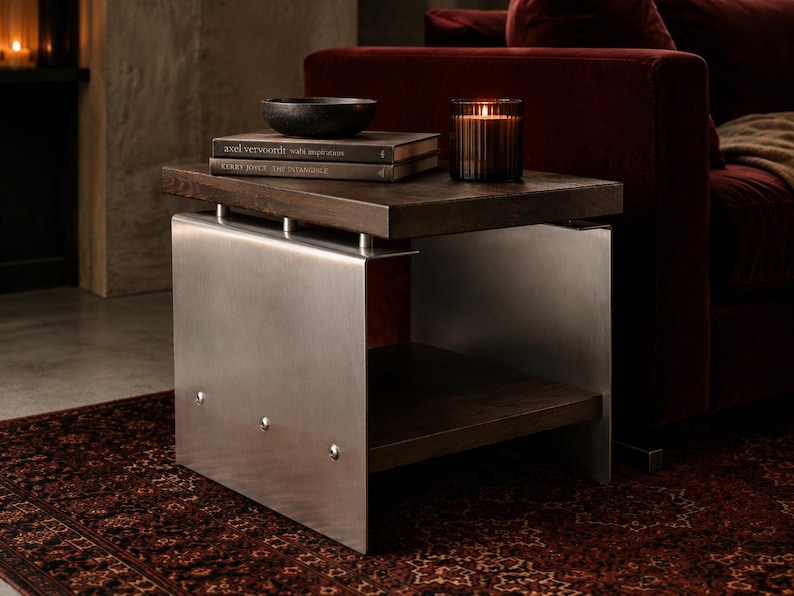 Op de afbeelding: Een bijzettafel met een donkerbruin houten blad en een onderste plank, ondersteund door twee geborstelde zilveren metalen zijpanelen die zijn bevestigd met zichtbare klinknagels. De tafel bevat twee gestapelde boeken en een glazen kaarshouder.