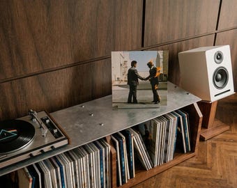 Op de afbeelding: Een houten opbergmeubel voor platen met een metalen bovenblad, waarop een platenspeler met een zwarte vinylplaat, een Pink Floyd Wish You Were Here albumhoes en een witte luidspreker staan. Het meubel bevat rijen vinylplaten in hoezen.