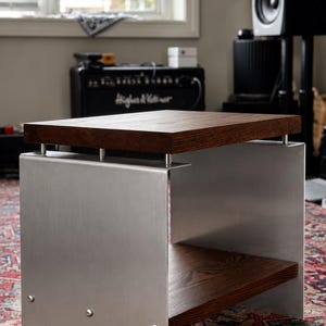 Op de afbeelding: Een bijzettafel met een dik, donkerbruin houten blad en een bijpassende onderste plank, ondersteund door twee verticale zijpanelen van geborsteld zilverkleurig metaal. De metalen panelen zijn bevestigd met zichtbare zilveren bouten.