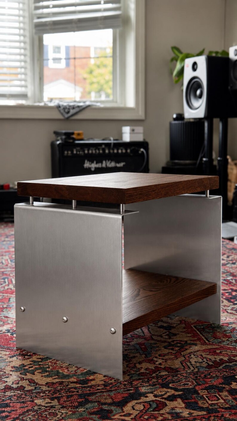 Op de afbeelding: Een bijzettafel met een dik, donkerbruin houten blad en een bijpassende onderste plank, ondersteund door twee verticale zijpanelen van geborsteld zilverkleurig metaal. De metalen panelen zijn bevestigd met zichtbare zilveren bouten.