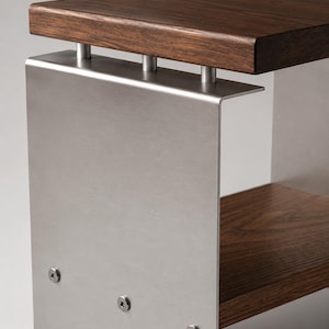 Op de afbeelding: Een bijzettafel met een donkerbruin houten blad en een onderste houten plank, verbonden door een frame van geborsteld zilverkleurig metaal. Het metalen zijpaneel is aan de houten onderdelen bevestigd met drie zichtbare ronde schroeven.