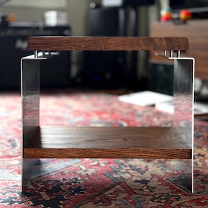 Op de afbeelding: Een kleine bijzettafel met twee horizontale planken van donkerbruin hout, verbonden door twee verticale zijpanelen van geborsteld metaal. De bovenste plank is met kleine zilverkleurige bevestigingen aan de metalen steunen vastgemaakt.