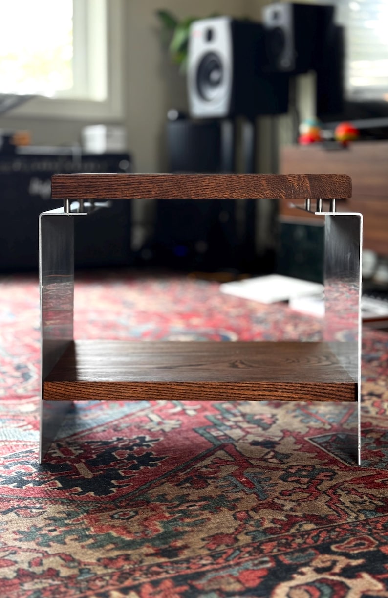 Op de afbeelding: Een kleine bijzettafel met twee horizontale planken van donkerbruin hout, verbonden door twee verticale zijpanelen van geborsteld metaal. De bovenste plank is met kleine zilverkleurige bevestigingen aan de metalen steunen vastgemaakt.