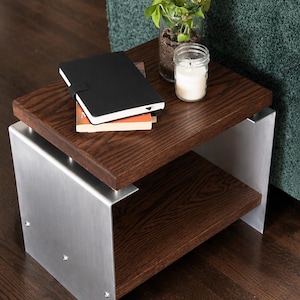 Op de afbeelding: Een bijzettafel met twee donkerbruine houten planken ondersteund door twee zijpanelen van geborsteld zilverkleurig metaal. Op de bovenste plank liggen een zwart notitieboek, een kleine groene plant in een pot en een witte kaars in een pot.