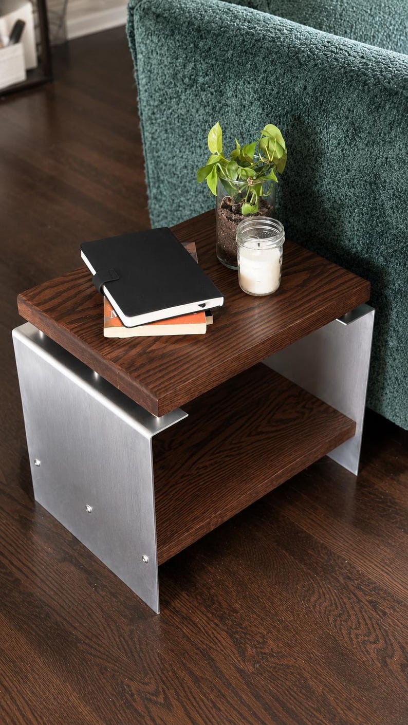 Op de afbeelding: Een bijzettafel met twee donkerbruine houten planken ondersteund door twee zijpanelen van geborsteld zilverkleurig metaal. Op de bovenste plank liggen een zwart notitieboek, een kleine groene plant in een pot en een witte kaars in een pot.