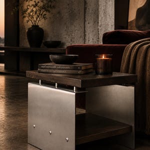 Op de afbeelding: Een vierkante bijzettafel met een donkerbruin houten blad en een onderste plank, ondersteund door twee geborstelde zilveren metalen zijpanelen vastgezet met zichtbare klinknagels. De tafel bevat twee boeken, een donkere kom en een glazen kaarshouder.