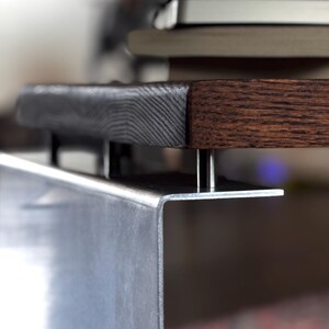 Op de afbeelding: Een donkerbruin houten tafelblad met een zichtbare houtnerfstructuur wordt ondersteund door een zilverkleurige metalen beugel. Het metalen onderdeel heeft een scherpe haakse bocht en is met kleine metalen afstandhouders aan de onderkant bevestigd.