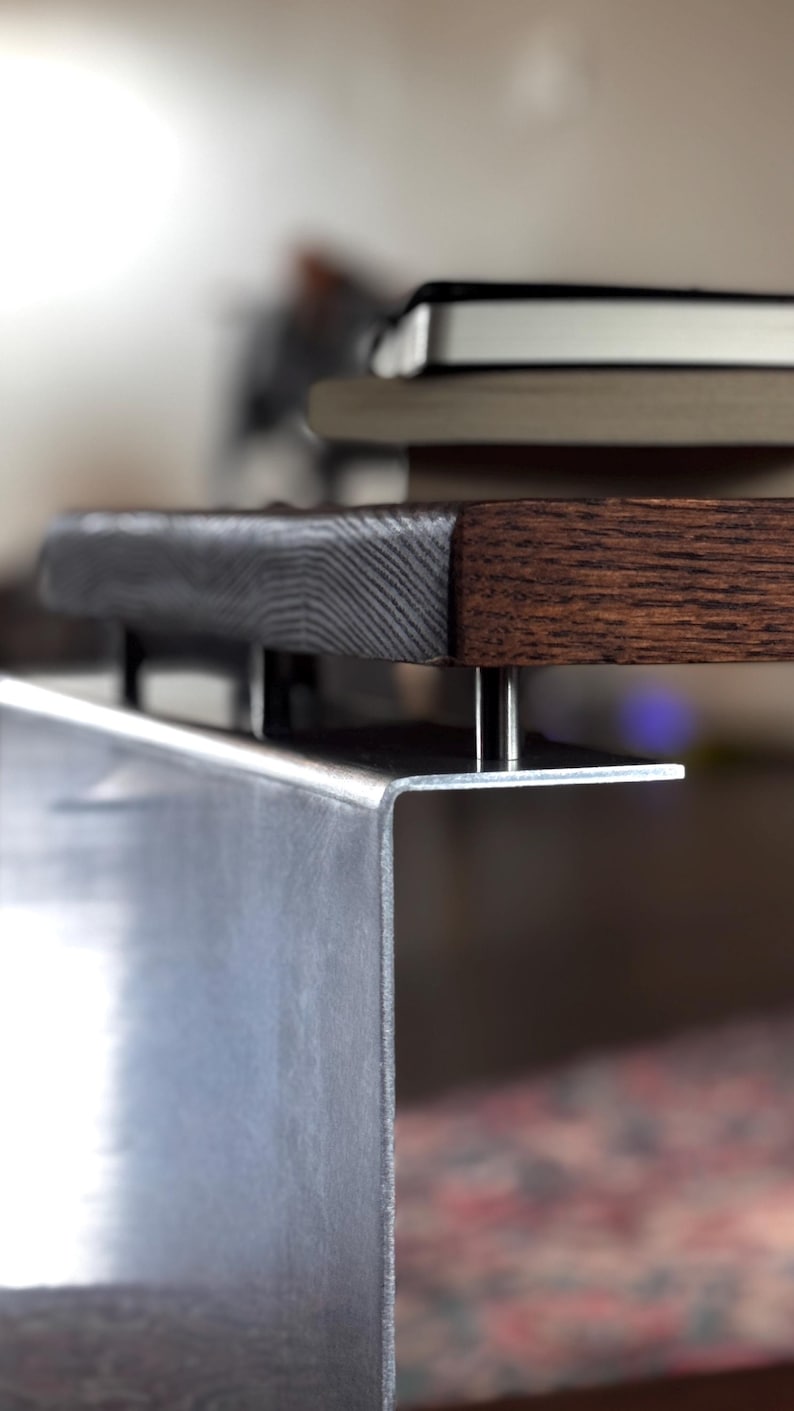 Op de afbeelding: Een donkerbruin houten tafelblad met een zichtbare houtnerfstructuur wordt ondersteund door een zilverkleurige metalen beugel. Het metalen onderdeel heeft een scherpe haakse bocht en is met kleine metalen afstandhouders aan de onderkant bevestigd.
