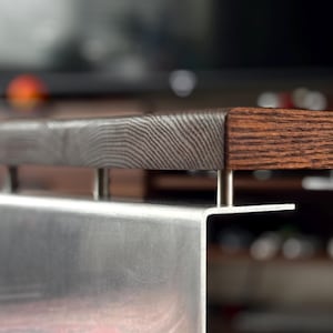 Op de afbeelding: Een meubelstuk met een dik, donkerbruin houten blad met zichtbare houtnerven. Het houten oppervlak is verhoogd boven een geborsteld zilveren metalen voet door vier kleine, cilindrische metalen afstandhouders die de twee materialen verbinden.
