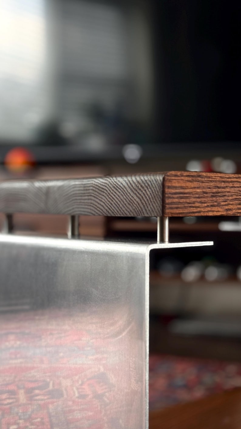 Op de afbeelding: Een meubelstuk met een dik, donkerbruin houten blad met zichtbare houtnerven. Het houten oppervlak is verhoogd boven een geborsteld zilveren metalen voet door vier kleine, cilindrische metalen afstandhouders die de twee materialen verbinden.